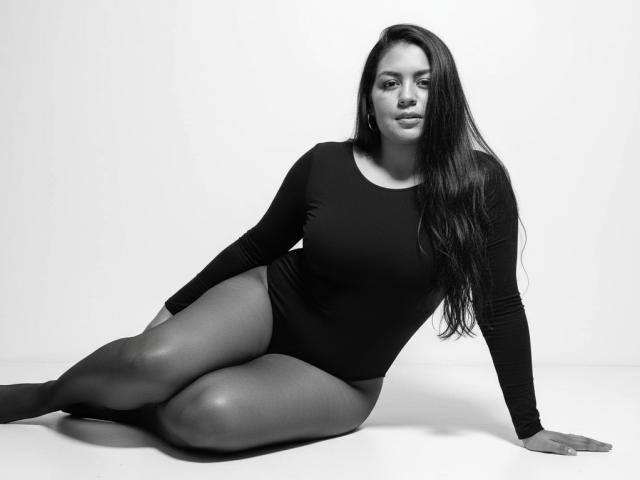AthenaTheas has long dark hair, wearing a black long-sleeve bodysuit, sitting with legs crossed, showing her ass, in a black-and-white photo. She's looking at the camera.