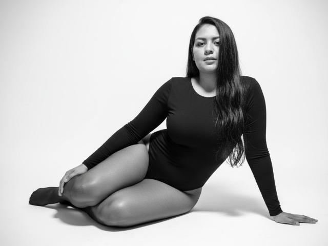 AthenaTheas is sitting with legs folded, wearing a black long-sleeve leotard, showing her tits and ass. She has long dark hair, and a serious expression. Black-and-white photo.