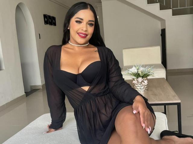 LanaRossel with black sheer dress, showing big boobs in a black bra, red lipstick, long black hair, choker, sitting, smiling, white chair, wooden table, white flowers, tan skin.