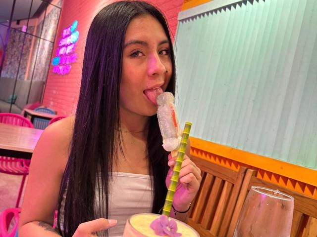 ChloeXk, avec de longs cheveux noirs, la langue tirée, léchant un bâtonnet de glace sucré vert, portant un débardeur blanc, assise à une table en bois..