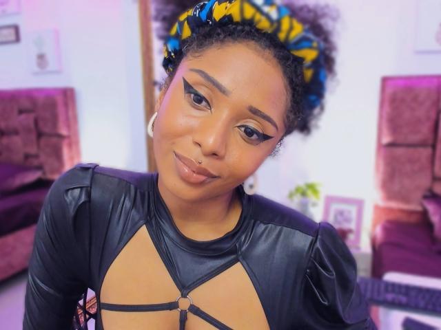 MeganWlliams has dark skin, curly black hair with blue and yellow beads, winged eyeliner, and a nose ring. She wears a black leather top with strappy details.