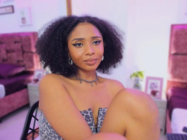 MeganWlliams with curly black hair, wearing a strapless patterned dress, sitting with knees up, choker necklace, and earrings, smiling slightly, in a softly lit room.