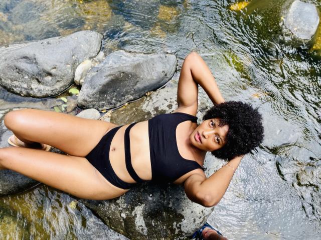 RoldanStefy with curly black hair, lying on rocks in a clear stream, wearing a black one-piece swimsuit, hands behind head, legs apart, displaying her ass and small tits.