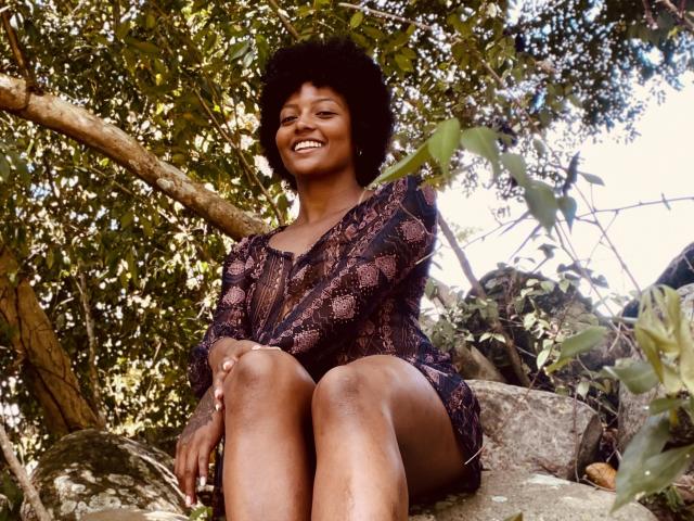 RoldanStefy, dark-skinned with afro, smiling, sitting on rocks, wearing a lacey, see-through purple top, showing her tits, in a sun-dappled forest.