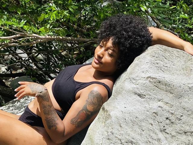 RoldanStefy with curly black hair, tan skin, and tattoos, lounging on a rock in a black bikini, eyes closed, smiling, sunlit, green foliage in the background.