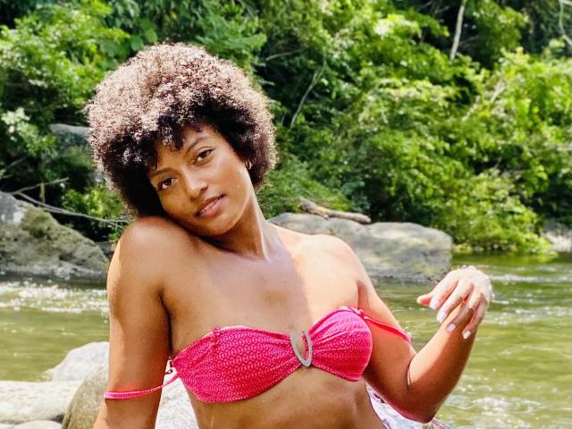 RoldanStefy with curly brown hair, brown skin, wearing a pink crocheted bikini top, showing small tits, by a green, rocky stream. She's looking at the camera, slightly tilted.