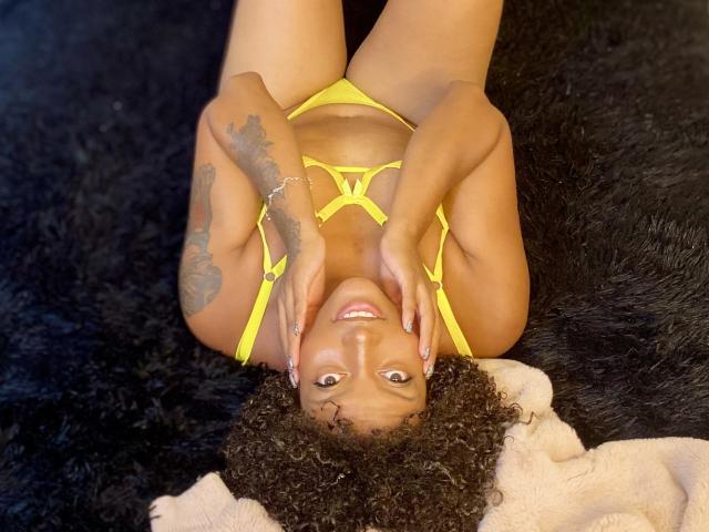 RoldanStefy, lying on a black furry surface, wearing a yellow strappy bikini, hands on cheeks, smiling, curly black hair, tattoo on right arm, cute, brown skin, small breasts.