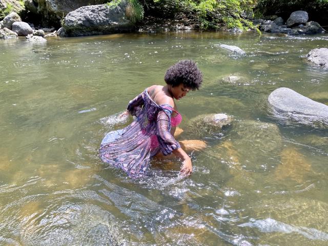 RoldanStefy with curly black hair, wearing a pink and purple dress, kneeling in a shallow, rocky stream, wet dress clinging to her bare ass. Water ripples around her.