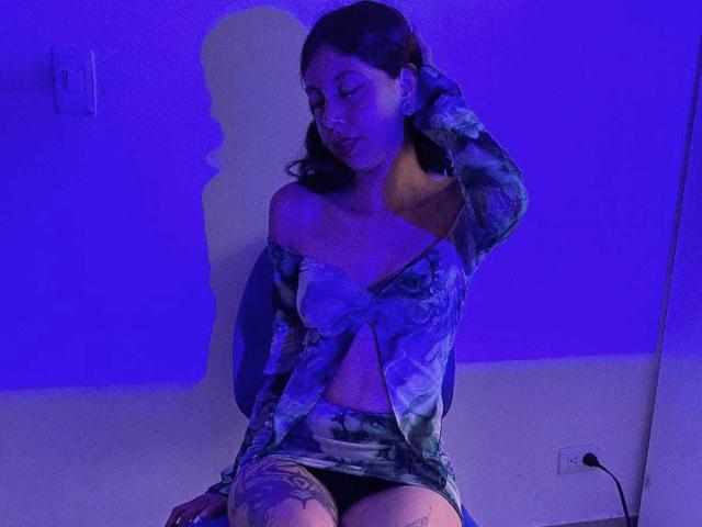 CamiVega with dark skin, wearing a blue and green floral off-shoulder dress, has a tattoo on her thigh, sitting, hand in hair, blue light, shadow on wall.