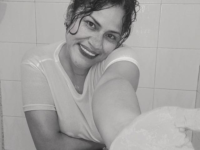 Grayscale photo of IvannaDuran with wet curly hair, smiling, wearing a wet white t-shirt, holding a soapy arm up, showing visible soapy bubbles, and a cute, freckled face.