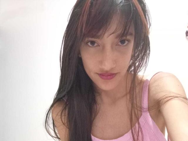 Kiriam Gomez, con cabello largo y castaño oscuro, un top rosa y lápiz labial rosa, mirando a la cámara con una ligera sonrisa, mostrando sus pequeños senos y su cara linda..