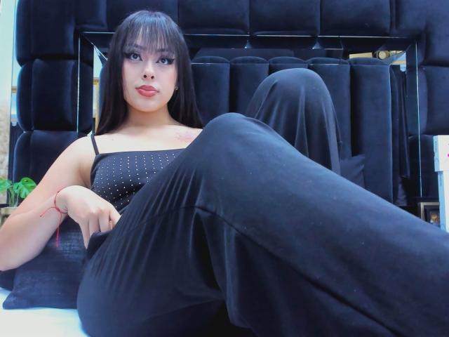 AlisonTatcher with long black hair, bangs, and full red lips, wearing a black sparkly top and black pants, sitting with legs up, showing her tits.