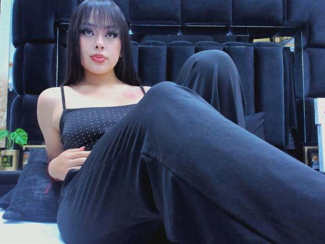 AlisonTatcher with long black hair, wearing a black sparkly top and black pants, sitting with legs crossed, showing small boobs and a tattoo on her right shoulder.