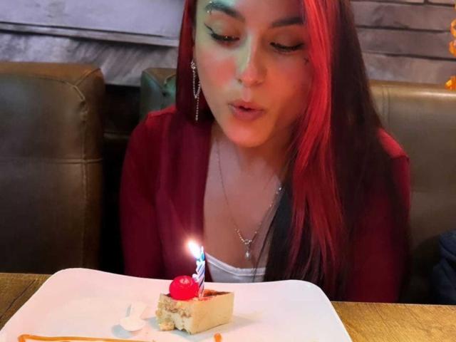 AmyStreetwise69 with red hair, looking down at a small cake with a lit candle, wearing a red jacket, in a brown leather booth. Cake has a cherry on top.