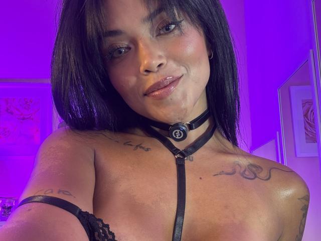 MajoJones with dark hair, glossy lips, wearing black harness, tattoos on chest and arm, purple lighting, smiling at camera. Boobs partially covered by lace.