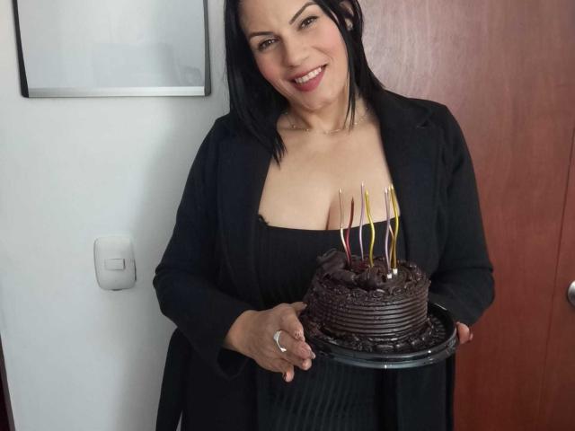 LizaRussell with black hair, smiling, wearing a black dress and blazer, holding a chocolate cake with candles, exposing her large boobs. She's standing indoors, holding the cake.