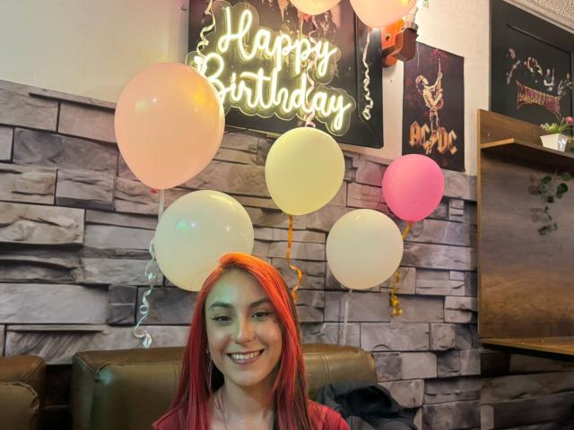 AmyStreetwise69 with red hair, smiling, has white, pink, and yellow balloons in a room with stone wall, neon "Happy Birthday" sign, and a poster in the background.