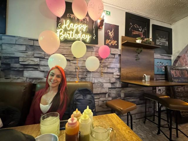 AmyStreetwise69 with red hair, tits visible, smiling at table with balloons, sauces, and drinks in a cozy cafe, stone wall and wooden furniture in the background.