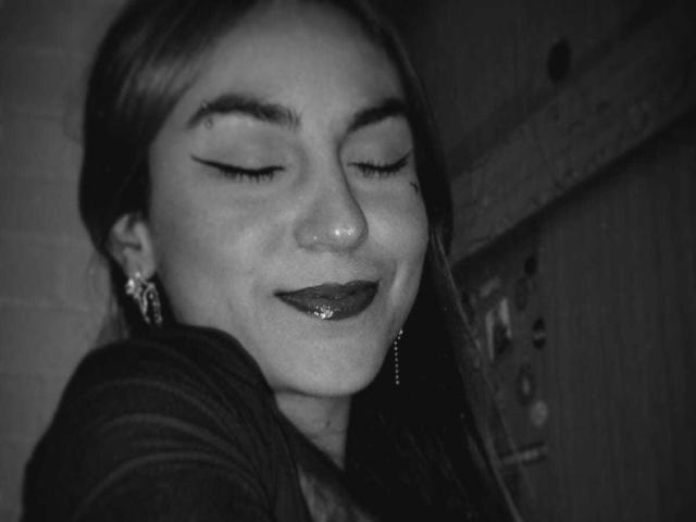 AmyStreetwise69 with closed eyes, smiling, wearing glittery lipstick, dark eyeliner, and dangling earrings, in a grayscale photo. Her nose is slightly shiny. She has long, straight hair.