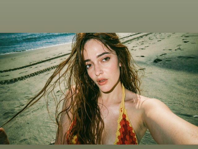 JaneRoa with wet, brown hair, wearing a red and yellow lace bikini top, showing her small tits, and a slightly open mouth, on a sandy beach with footprints.
