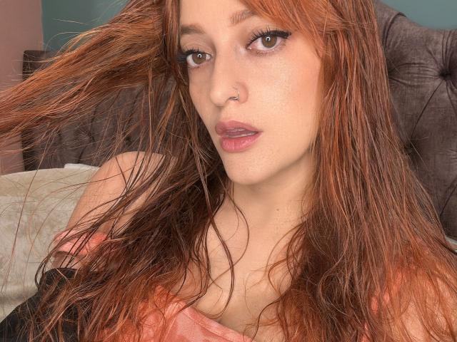 JaneRoa with wet, red-orange hair, nose piercing, brown eyes, and pink lips, wearing a pink top, showing her tits, looking into the camera.