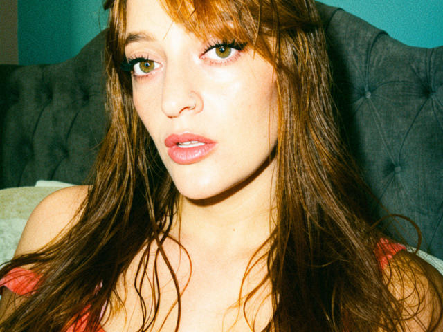 JaneRoa has long brown hair, green eyes, and pink lips. She's looking slightly to the side with a subtle, relaxed expression. She's wearing a red top.