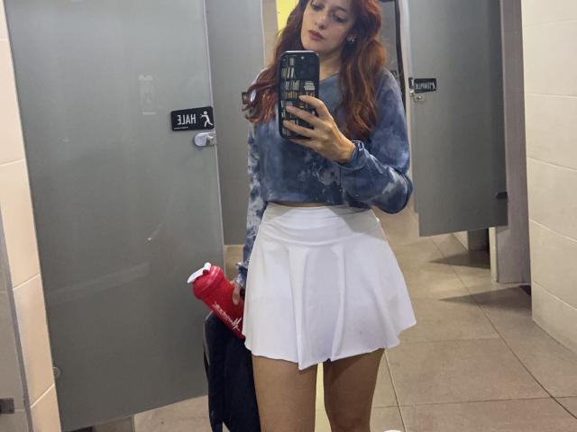 JaneRoa with red hair, wearing a blue tie-dye crop top and white pleated skirt, holding a red water bottle, standing in a bathroom, taking a mirror selfie.