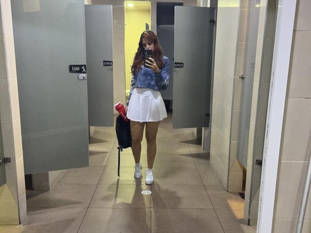 JaneRoa with long brown hair, wearing a blue hoodie, white skirt, and white sneakers, holding a red bag and black umbrella, taking a mirror selfie in a bathroom.