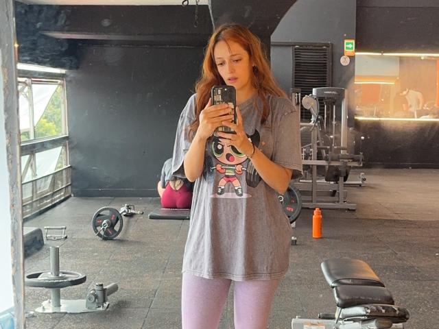 JaneRoa with long red hair, wearing a gray t-shirt with a cute print, pink leggings, taking a mirror selfie in a gym, holding a smartphone, standing with a small ass.