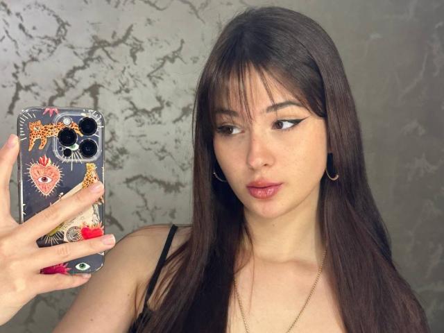 EmogeneBusalacchi with long brown hair, wearing a black top, holding a phone with colorful stickers, looking at the camera with winged eyeliner, and slightly parted pink lips.