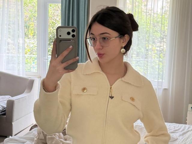 EmogeneBusalacchi with glasses, dark hair in a bun, white fluffy jacket, holding phone, taking selfie, looking at camera, small tits, fair skin, bedroom, daytime, green curtain.