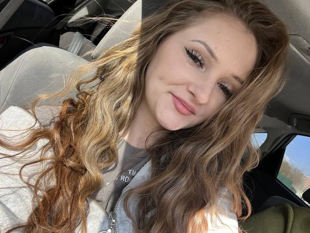 PamelaXu with wavy brown hair, thick eyelashes, and a slight smile, wearing a grey top, sitting in a car. Her tits are not visible.