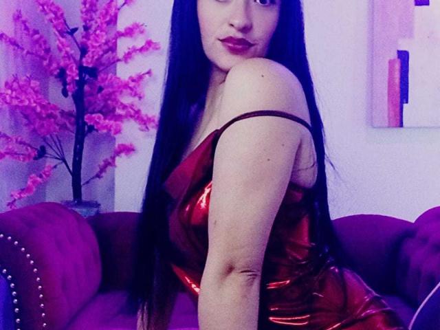 Alanna Scott mit langem, schwarzem Haar, einem roten, glänzenden Kleid, freiem Schulterteil, sitzend auf einem violetten Sofa, die über ihre Schulter schaut, mit rotem Lippenstift, violettes Licht. Brüste sind unter dem Kleid sichtbar..