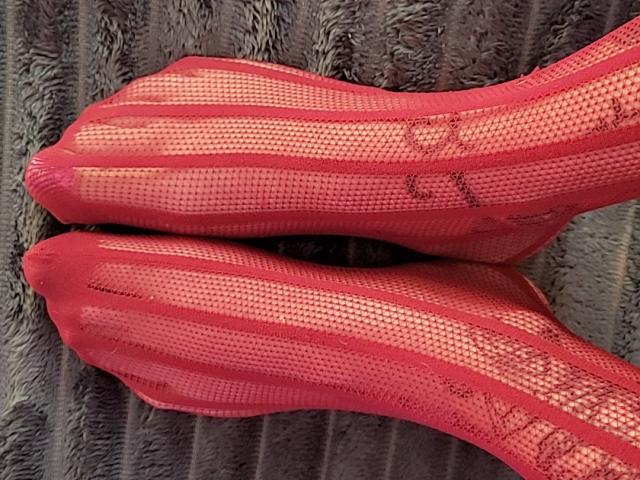 Les pieds d'ElixirShanna, chaussés de bas résille rouges et rayés, montrant une légère usure, sont croisés à la cheville. La texture est en maille avec des lignes visibles..