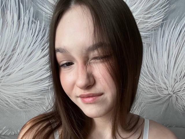 AnitraAmphy has fair skin, brown straight hair, and is winking with a small smile. She's wearing a white tank top and has subtle makeup. Her tits are small.