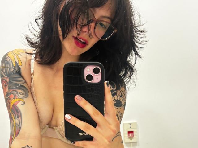 IsabellaArias with wavy black hair, glasses, red lipstick, and tattoos, taking a mirror selfie with her phone, showing her boobs in a pink bra, white wall in background.