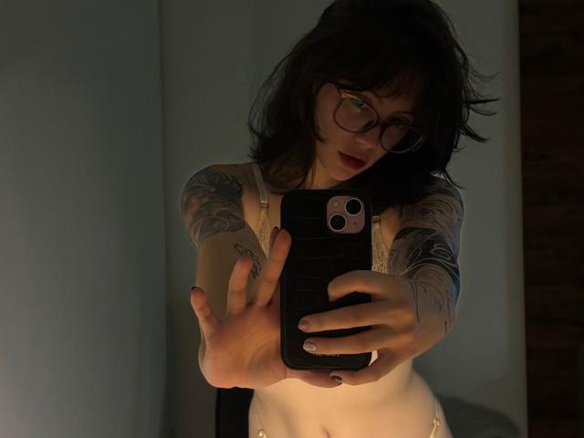 IsabellaArias with tattoos, glasses, and dark hair, taking a mirror selfie with her phone, extending her arm, showing her tits in white lingerie, in dim light.