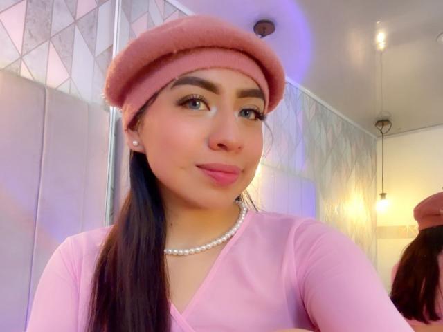 ZoeRedds trägt eine rosa Beere, ein pinkes Langarmshirt und eine Perlenkette, mit pinkem Lippenstift und langen braunen Haaren, und lächelt in einem Spiegel-Selfie..