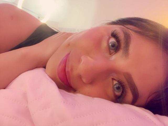 ZoeRedds with pink lips and blue eyes, lying on a pink pillow, wearing a black top, has a small mole on her right cheek. Her eyelashes are long and dark.