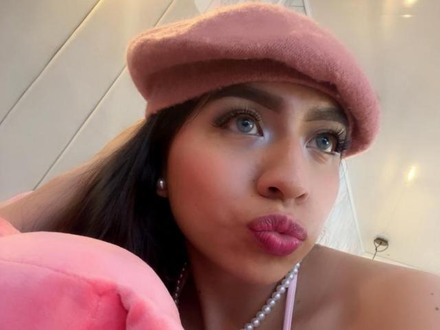 ZoeRedds has pink lips, blue eyes, and dark hair, wearing a pink beret and pearl necklace, with a pink blanket in the foreground, making a pouty face.