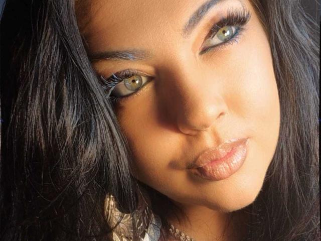 SatisfyMyDesires has dark, wavy hair, green eyes, and glossy, pouty lips. She's looking at the camera with a slightly tilted head, giving a sultry, sexy look.