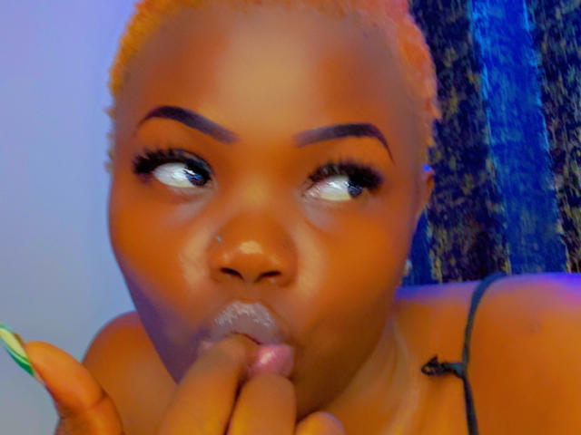 RealNasty69 with orange hair, dark eyebrows, and a nose ring, sucking on a finger with glossy lips, wearing a spaghetti strap top, blue and purple lighting.