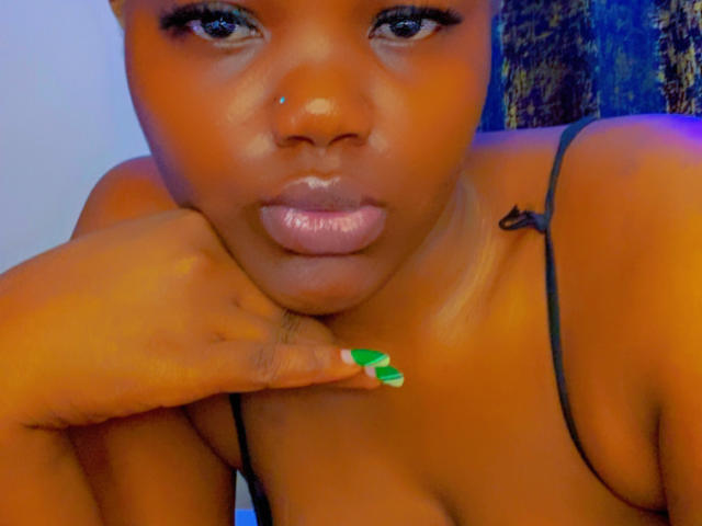 RealNasty69's brown face, shiny pink lips, green nail polish, black spaghetti strap, nose piercing, and hand resting on chin in a colorful, tight shot.