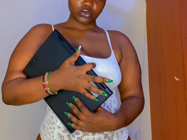 RealNasty69 with dark skin, wearing a white lace tank top and white lace underwear, holding a black tablet, green nails, and colorful bracelets, looking serious.