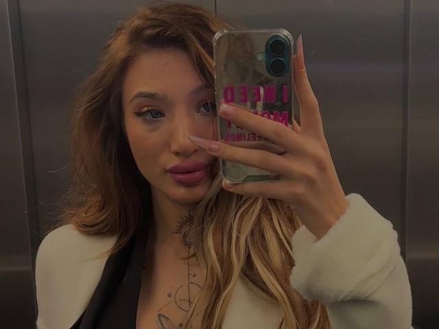 SiennaJayden with long, wavy blonde hair, wearing a white jacket, taking a mirror selfie in an elevator, showing a tattoo on her chest, holding a phone with pink text.