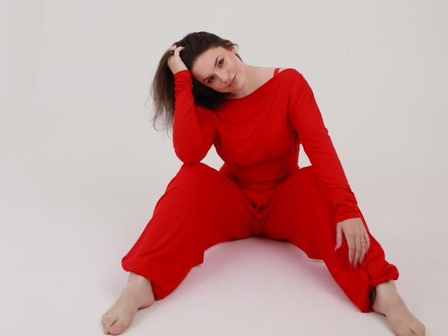 Dreia, sitting on the floor, wearing a red long-sleeve shirt and red pants, legs spread, hand in brown hair, bare feet, red outfit, white background, soft smile.