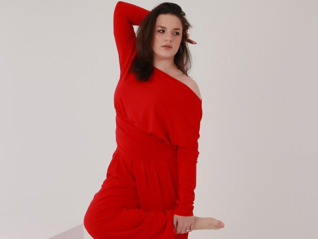 Dreia with pale skin and dark brown hair, wearing a red, off-shoulder dress, has one arm raised, showing her tits, and sits with one leg up.