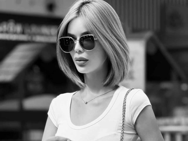 Fotografía en blanco y negro de HotCumAdriana69x con un corte de pelo bob, usando gafas de sol oscuras, una camiseta blanca y un bolso de cadena, mirando ligeramente hacia un lado..
