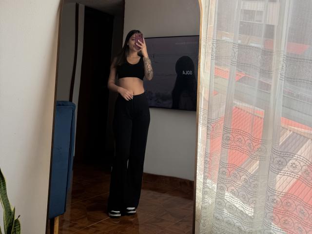ThaliaAres with tattoos, wearing black sports bra and high-waisted black pants, taking a mirror selfie, showing her flat stomach and black-and-white sneakers.
