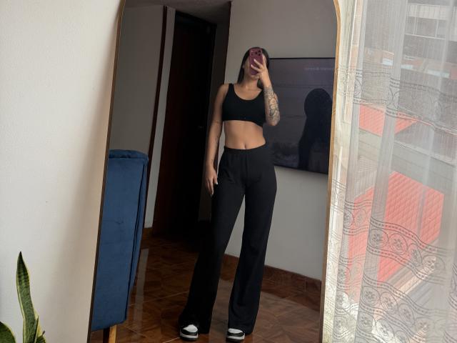 ThaliaAres with tattoos, black sports bra, black pants, and white shoes, standing in front of a mirror, holding a phone, showing her flat stomach and small ass.
